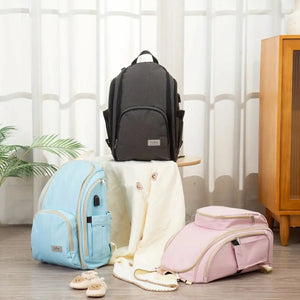 Baby diaper backpacks in dark grey, sky blue and pink colors displayed in a nursery setting