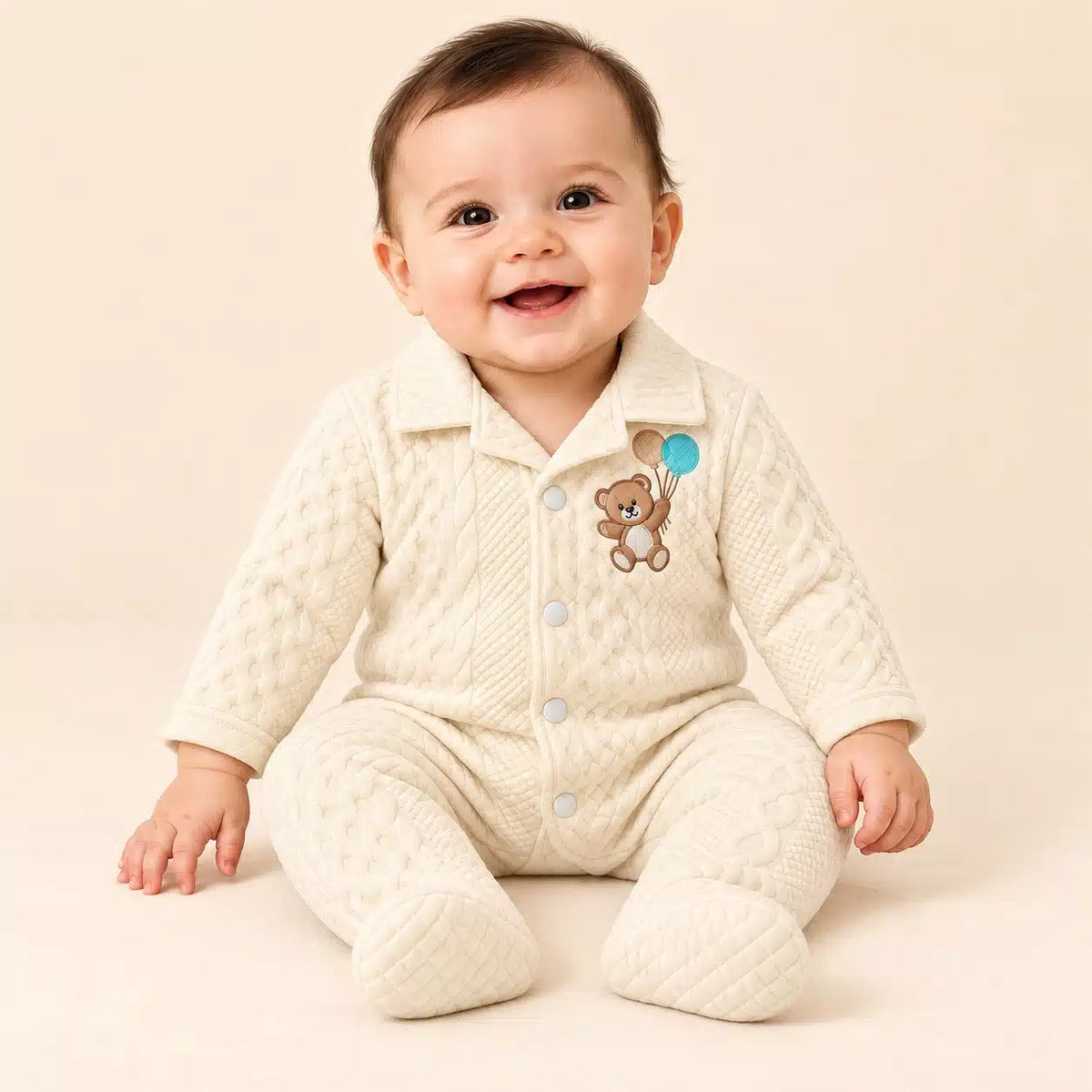 Baby wearing cream winter quilted footed romper with teddy bear embroidery in seated pose