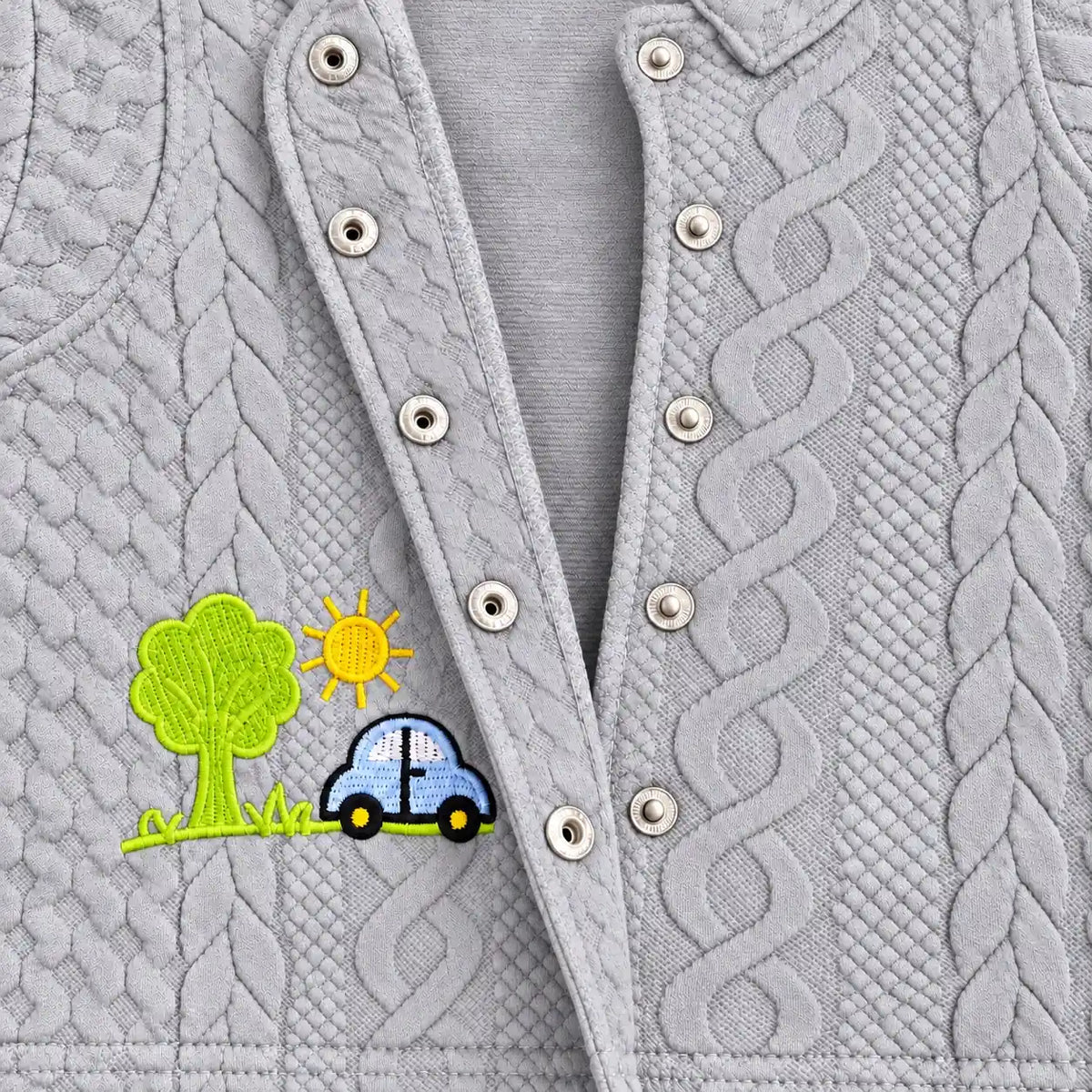 Close-up of open metal snap buttons, raised cable-style texture and car embroidery on light grey baby shirt