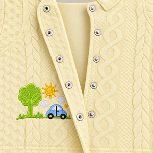 Close-up of open metal snap buttons, raised cable-style texture and car embroidery on cream baby shirt